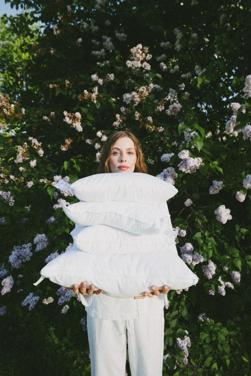 A person holding white Snoooze pillows in front of lilac flowers.